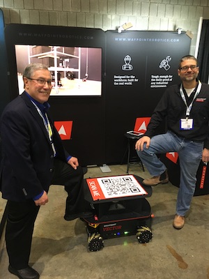 Numina集团首席执行官Dan Hanrahan(左)和Waypoint机器人公司的Jason Walker与Waypoint公司的AMRs之一。AMR的“Kingpin”模块(在背景显示屏上使用)可以移动重型货架或手提袋/箱子，允许灵活的订单挑选